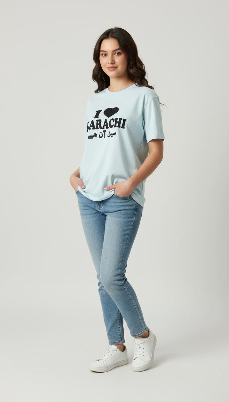 A woman stands against a light grey background, smiling with her hands in her pockets, wearing the Everyday Ease Tee—a soft, breathable I ♥ Karachi shirt in light blue with English and Urdu text—paired with blue jeans and white sneakers.