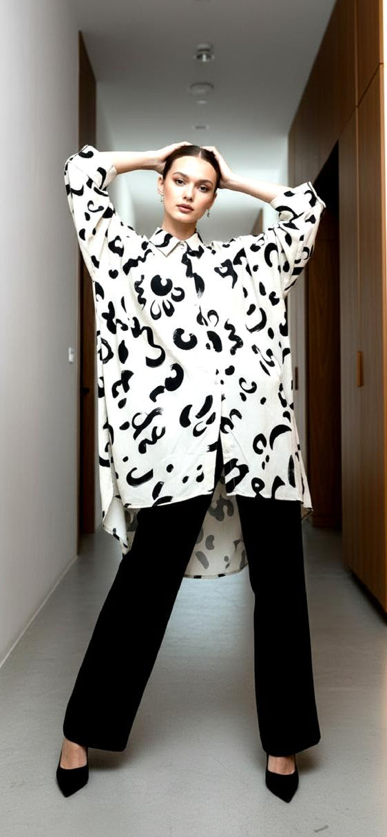 A woman stands confidently in a hallway, wearing the Chic Contemporary Fit Top—an oversized white shirt with black abstract patterns—paired with black wide-leg pants and heels, her hands resting on her head.