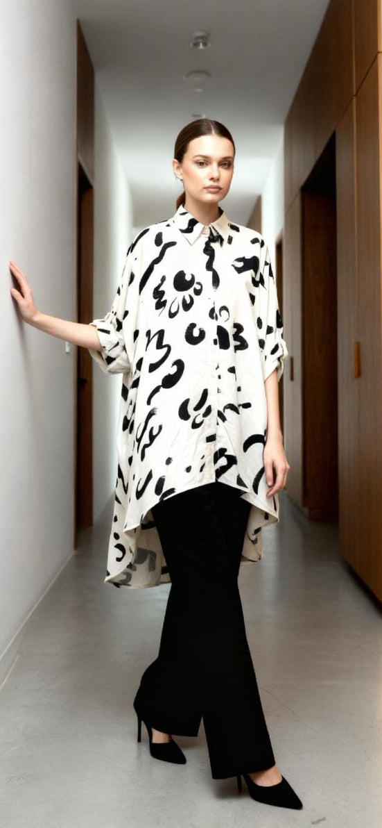 A woman stands in a modern hallway wearing the Chic Contemporary Fit Top—an effortlessly elegant loose white shirt with black abstract patterns—paired with black pants and heels as she rests one hand on the wall.