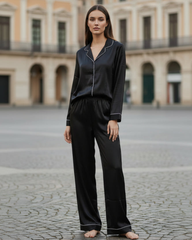 Woman wearing a black satin pajama set standing on a stone pavement with classical architecture in the background.
