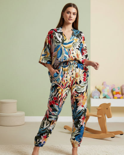 A woman stands barefoot in a room, wearing the Minimal Luxe Co-Ord—a colorful, floral-patterned set exuding minimal charm and understated elegance. Behind her are a wooden rocking horse, plush toys, and a green-beige wall.