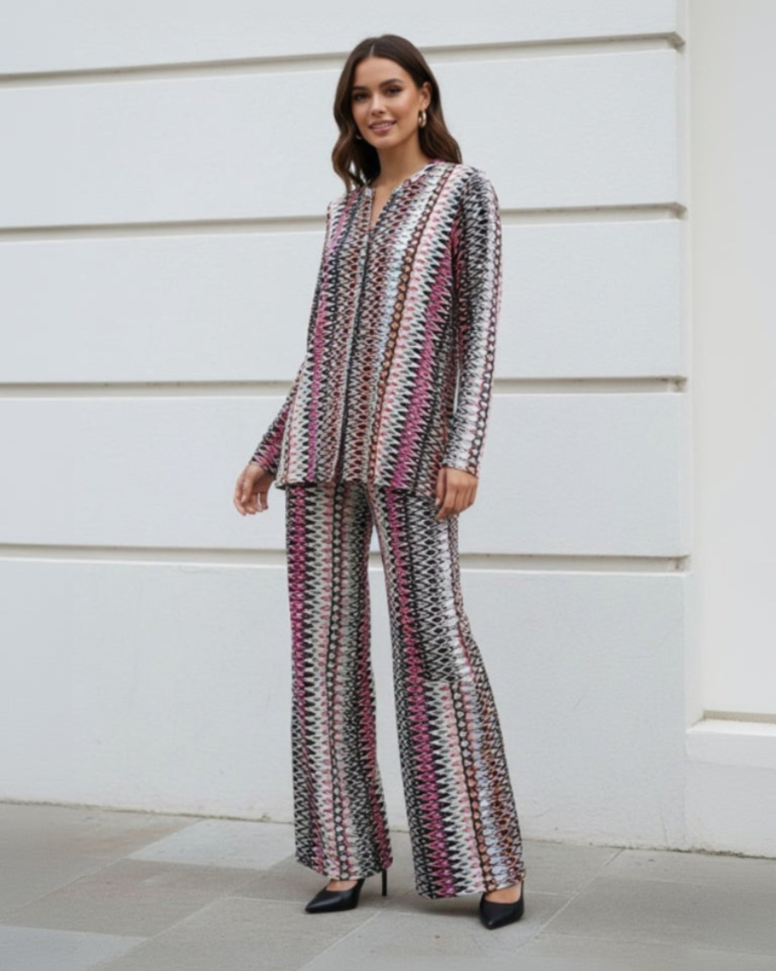 A woman with long brown hair smiles outdoors, wearing the Refined Flow Co-Ord—a stylish long-sleeved top and flared pants set with a colorful geometric pattern in pink, purple, black, and white. She pairs it with black heels.