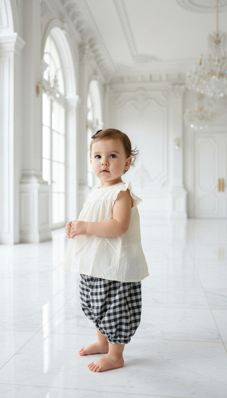 Child wearing a white sleeveless top and black checkered pants in a bright, white room.