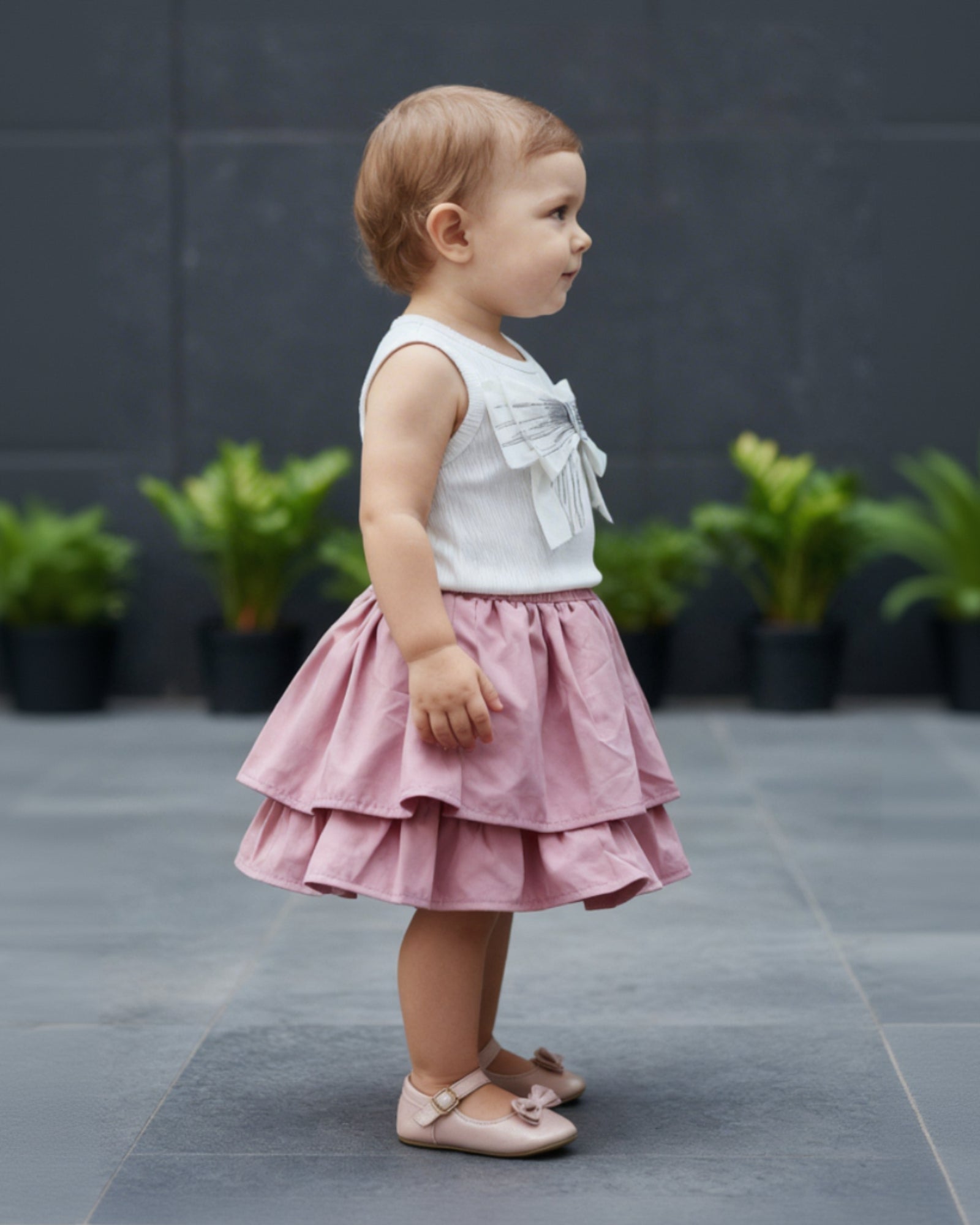 Child wearing a white top with bow details and a pink skirt standing on a gray pavement.