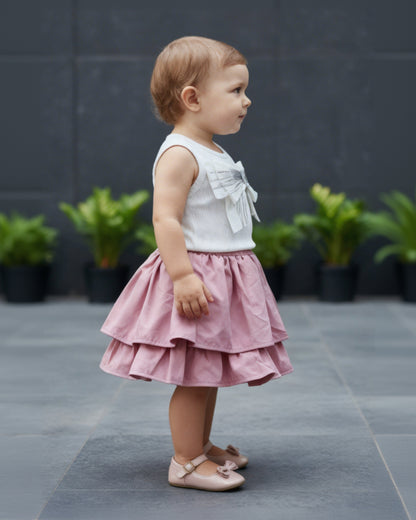 Child wearing a white top with bow details and a pink skirt standing on a gray pavement.