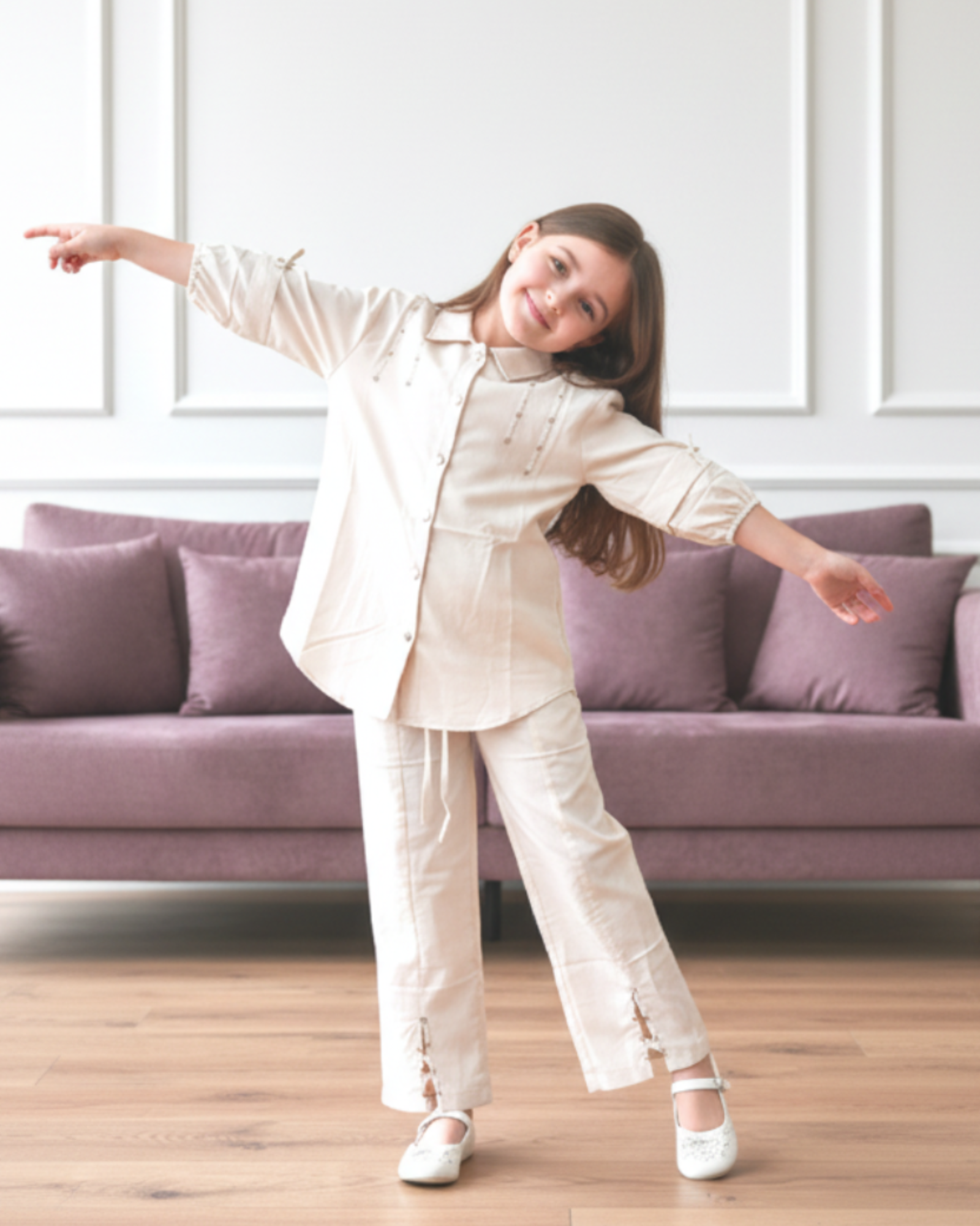 Child in a white outfit standing in a room with a purple couch and a framed picture on the wall.
