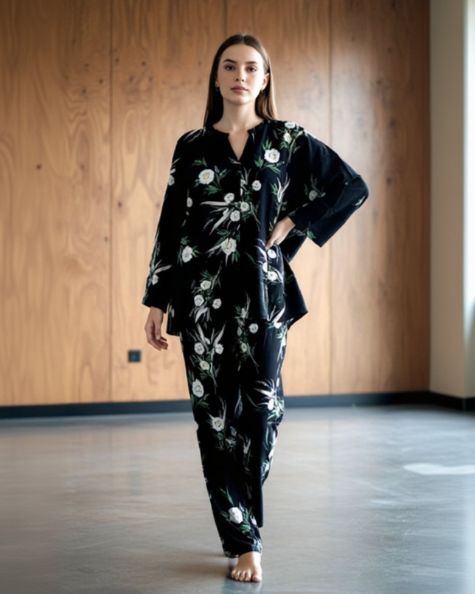 A woman with long brown hair walks barefoot indoors, wearing the easy-to-style Modern Muse Co-Ord in black floral, posing before a wooden wall for a contemporary look.