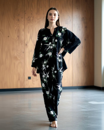 A woman with long brown hair walks barefoot indoors, wearing the easy-to-style Modern Muse Co-Ord in black floral, posing before a wooden wall for a contemporary look.