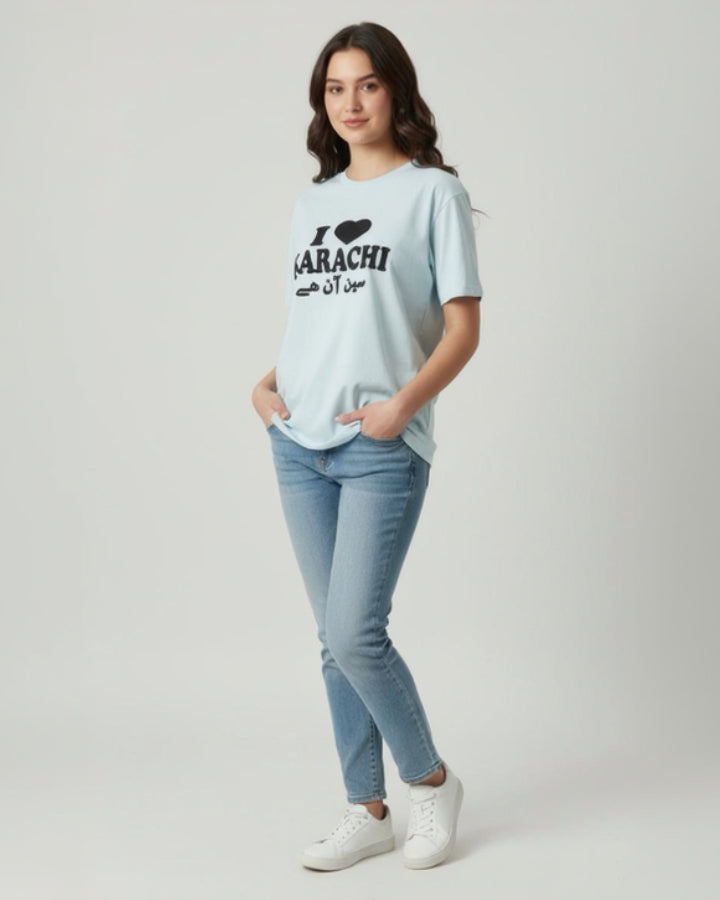 A woman stands against a plain backdrop, smiling with her hands in her pockets and wearing the Everyday Ease Tee—a soft, breathable shirt that says I ♥ Karachi in English and Urdu—paired with blue jeans and white sneakers.