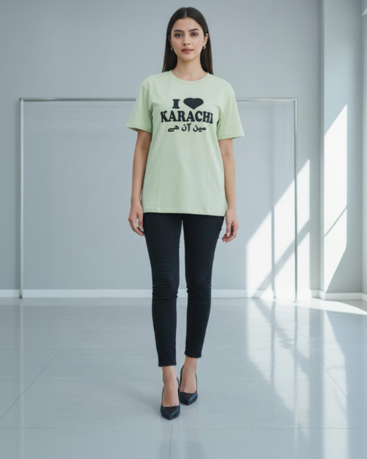Woman wearing a light green 'IKARACHI' t-shirt and black pants in a minimalistic setting.