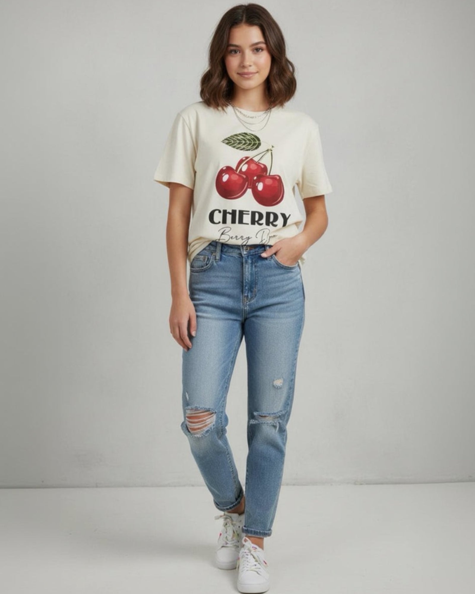 A young woman with shoulder-length brown hair stands against a plain background, wearing the Pure Comfort Tee featuring two red cherries and "CHERRY," teamed with blue ripped jeans and white sneakers for everyday comfort.