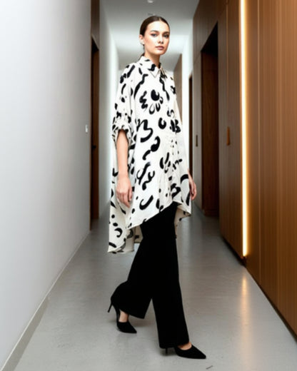A woman with slicked-back hair walks confidently down a modern hallway, showcasing understated elegance in the Chic Contemporary Fit Top—loose and timeless with bold black abstract patterns—paired with wide-leg black pants and sleek heels.