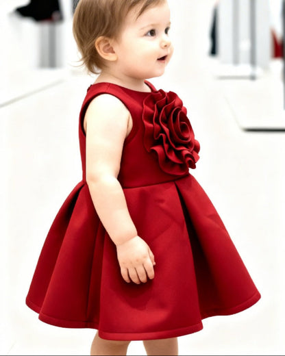 Child wearing a red dress with a large flower detail in a white room.