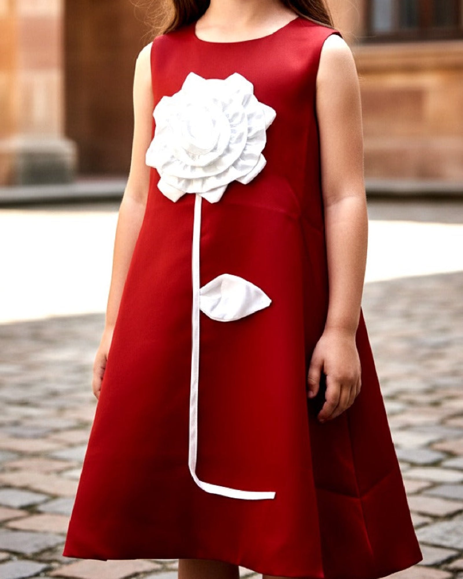 Young girl wearing a red dress with a white flower design in an outdoor setting.
