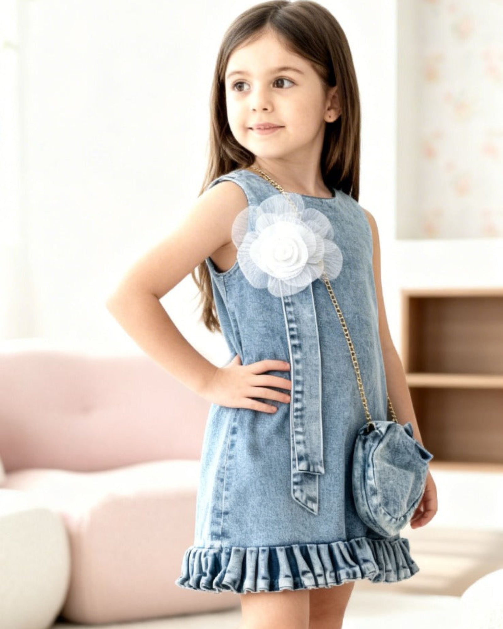 Young girl wearing a denim dress with a white flower accessory indoors.