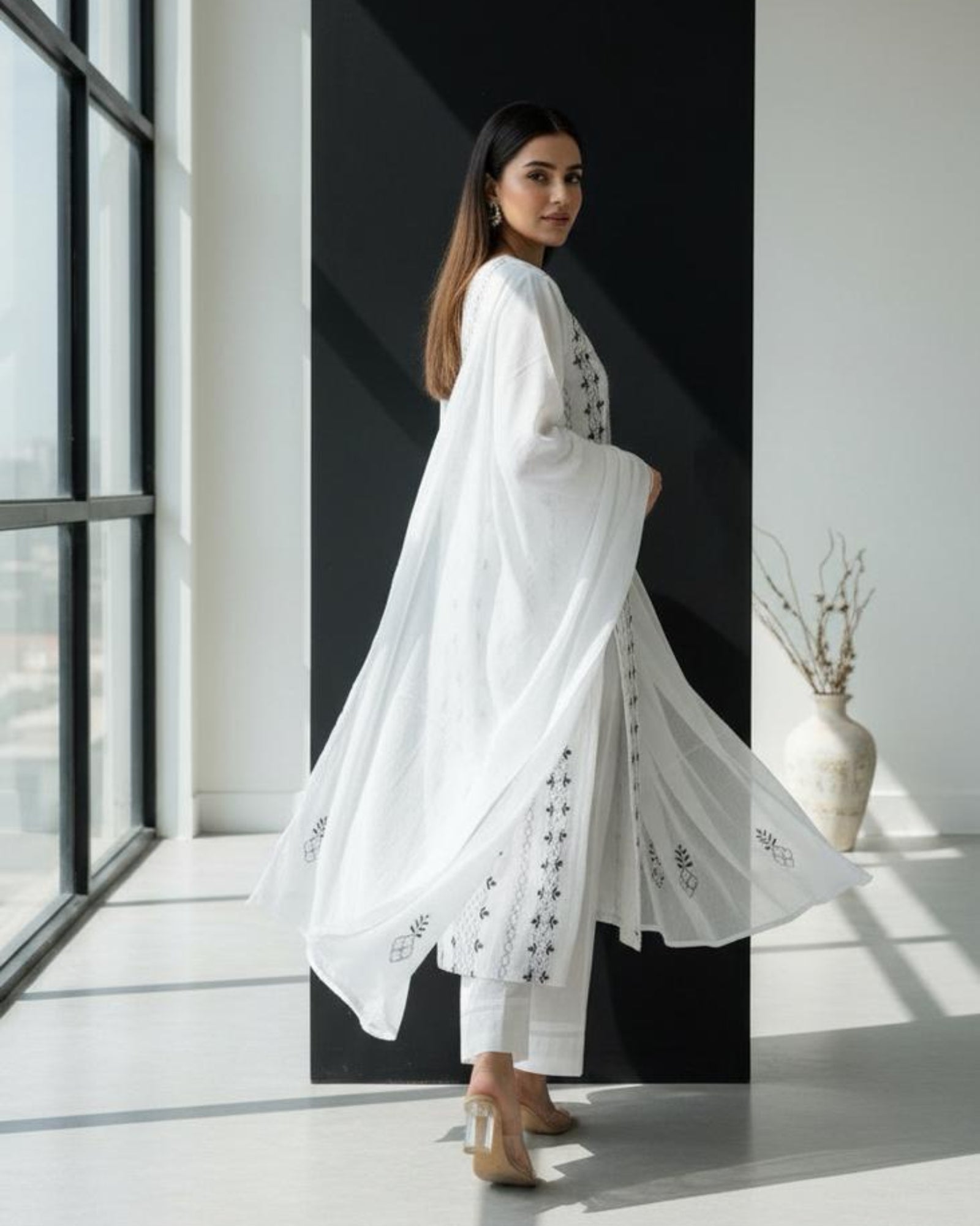 Woman in a white outfit standing in a modern interior setting with large windows.